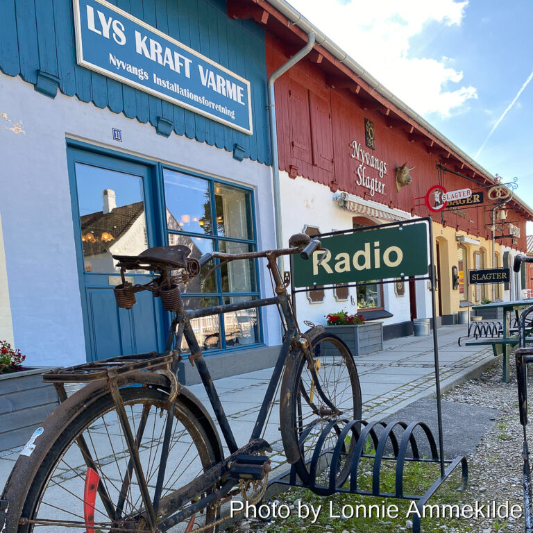 En gammel stort cykel står i et stor cykelstativ, på cukelstativet er der en mørkegrøn plade hvor der står Radio med gult. I baggrunden ses et blåt skilt med hvid kant hvor der står Lys Kraft Varme men hvidt