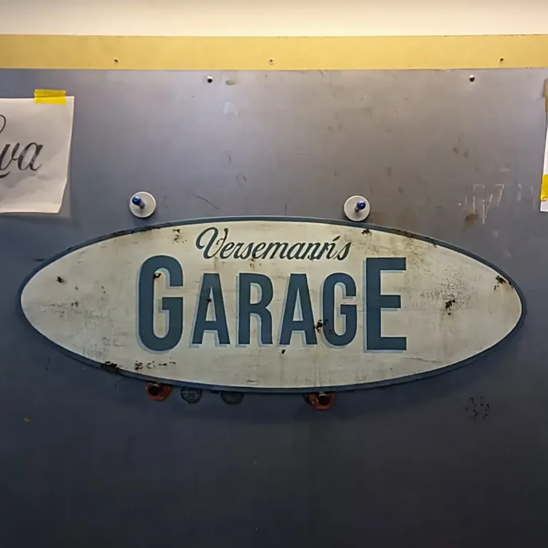 Ovalt skilt i lyseblå men en mørkeblå kant. Teksten på billedet er Versemanns Garage ig teksten er i mørkeblå med en lysere blå skygge.
