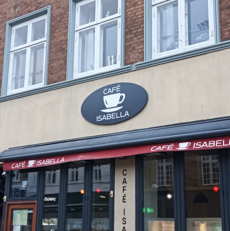Ellipseformet skilt i mørkegrå. Op skiltet står det Cafe øverst, under Cafe er er bollede af en stor kaffekop, og under koppen står det Isabella. Alt teksten på skiltet står med hvide bogstaver.