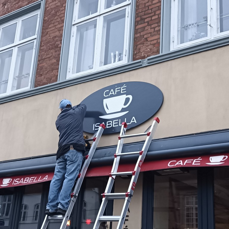 Mand står på stige og et ved at montere en ellipseformet skilt. Skiltet er gråt med teksten Cafe Isabella med et kaffekop i midten i hvidt.
