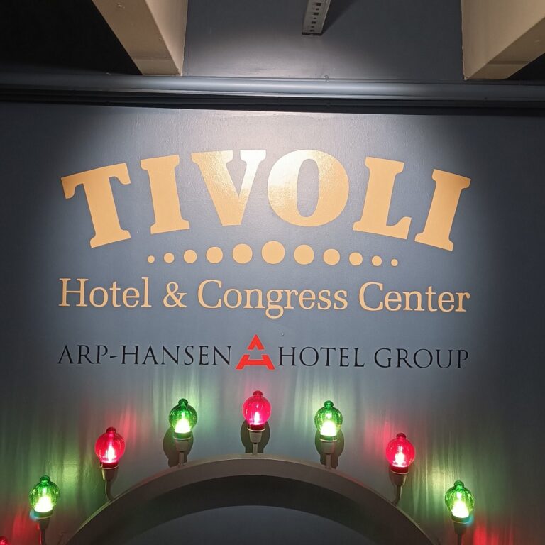 En blå væg, hvor der står med Tivoli Hotel og Congress Center med gul skrift. Undet den tekst står der ARP-Hansen Hotel Group med sorte bogstaver. Under alt teksten er der en grå bue hvor der er monteret røde og grønne lamper.