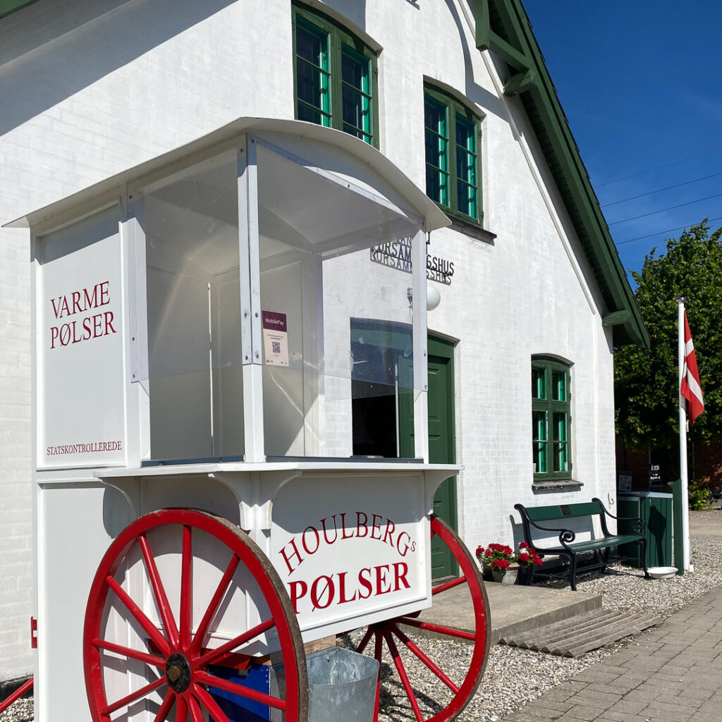 Håndmalet skilt på klassisk Houlberg pølsevogn med røde hjul foran historisk bygning.